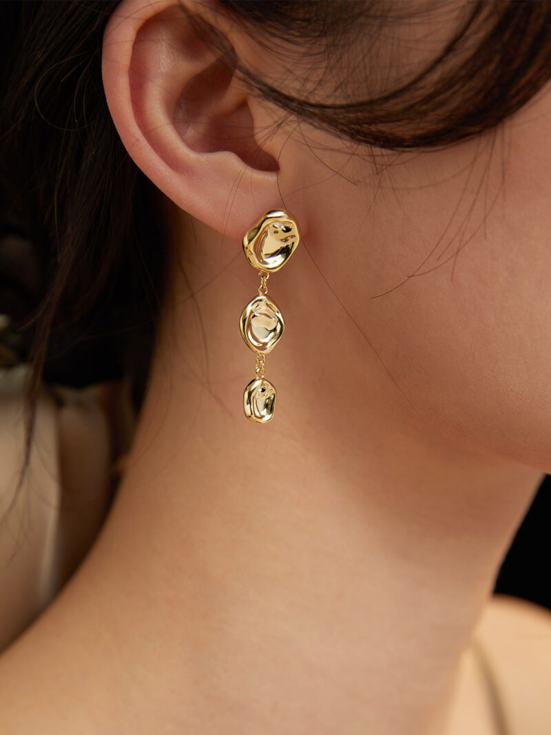 Close-up of a woman's ear wearing a long, gold, circular-link earring featuring a freshwater pearl with a focus on the jewelry against a blurred beige background. Elegant pearl jewelry