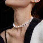 A close-up side view of a woman's neck adorned with a pearls. She wears a gold earring and a black and white striped garment, with hair blowing gently. Elegant pearl jewelry