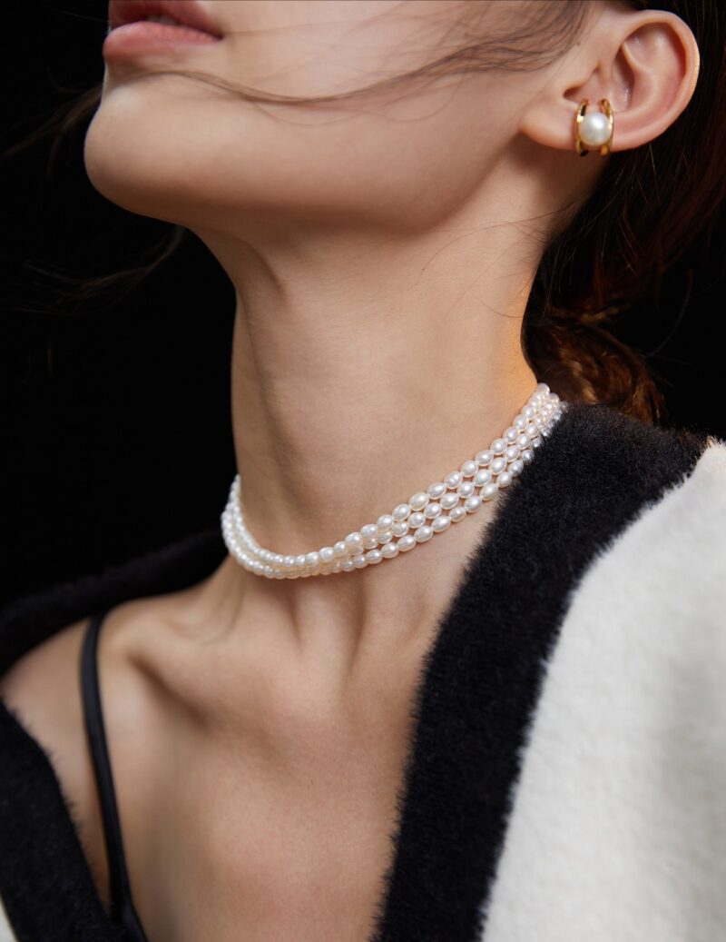 A close-up side view of a woman's neck adorned with a pearls. She wears a gold earring and a black and white striped garment, with hair blowing gently. Elegant pearl jewelry