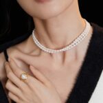 Close-up of a woman wearing jewelry, including a double strand  necklace and a large pearl ring, against a black background, focusing on the jewelry and lower part of her face. Elegant pearl jewelry