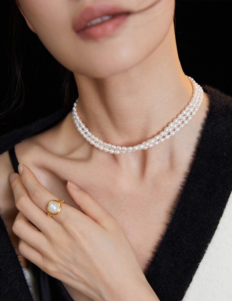 Close-up of a woman wearing jewelry, including a double strand  necklace and a large pearl ring, against a black background, focusing on the jewelry and lower part of her face. Elegant pearl jewelry