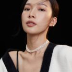 A portrait of an asian woman with a necklace and earrings, wearing a white and black blazer over a dark blouse, with her hair swept by a gentle breeze against a black background. Elegant pearl jewelry