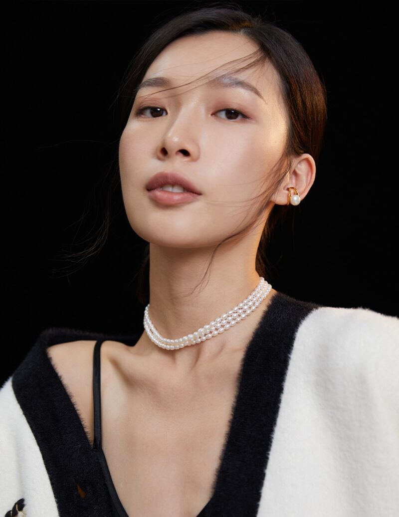 A portrait of an asian woman with a necklace and earrings, wearing a white and black blazer over a dark blouse, with her hair swept by a gentle breeze against a black background. Elegant pearl jewelry