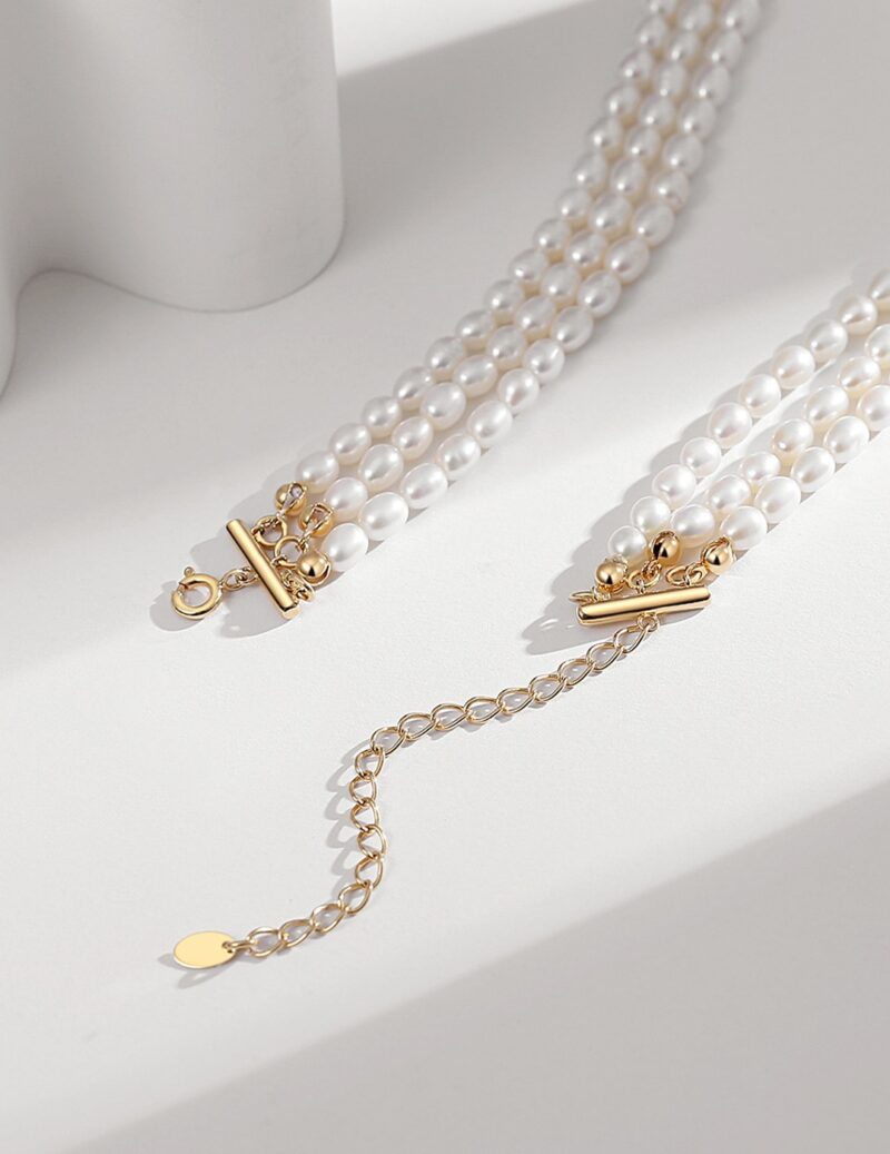 Two strands of pearls with golden clasps lie on a white surface, casting soft shadows in the sunlight, adjacent to a delicate gold chain with a small pendant. Elegant pearl jewelry