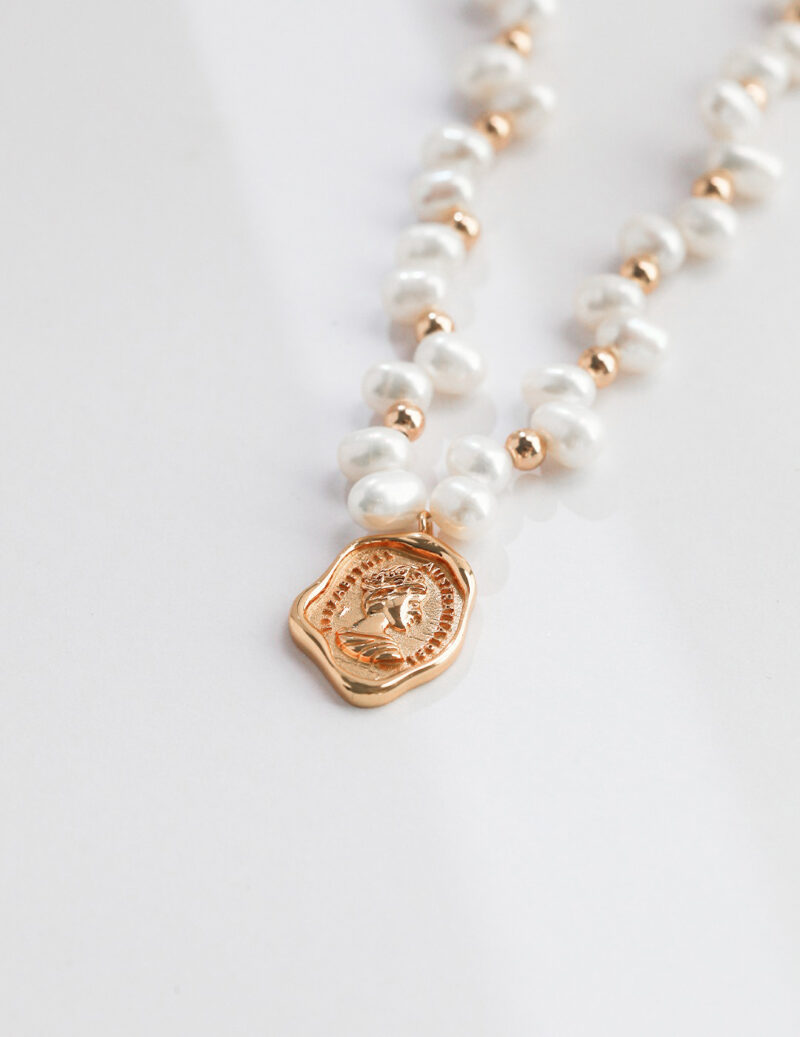 A necklace with gold beads and a gold pendant featuring an embossed design, resting on a plain white surface. Elegant pearl jewelry