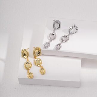 Two pairs of earrings displayed on a white tiered platform. Two pairs are interlocking circles in gold pearl jewelry, and one pair in sterling pearl jewelry, against a light gray background. Elegant pearl jewelry