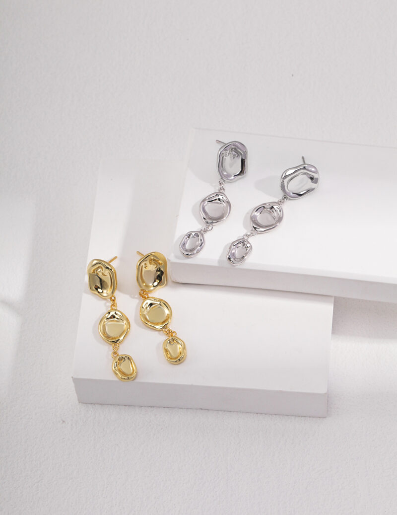 Two pairs of earrings displayed on a white tiered platform. Two pairs are interlocking circles in gold pearl jewelry, and one pair in sterling pearl jewelry, against a light gray background. Elegant pearl jewelry