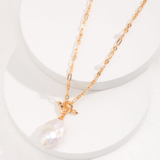 A delicate gold pearl jewelry necklace with a large, irregularly shaped freshwater pearl pendant, displayed on a cylindrical white stand with a textured surface. Elegant pearl jewelry