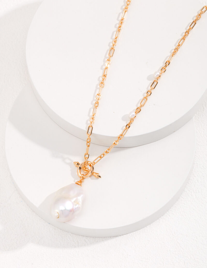 A delicate gold pearl jewelry necklace with a large, irregularly shaped freshwater pearl pendant, displayed on a cylindrical white stand with a textured surface. Elegant pearl jewelry