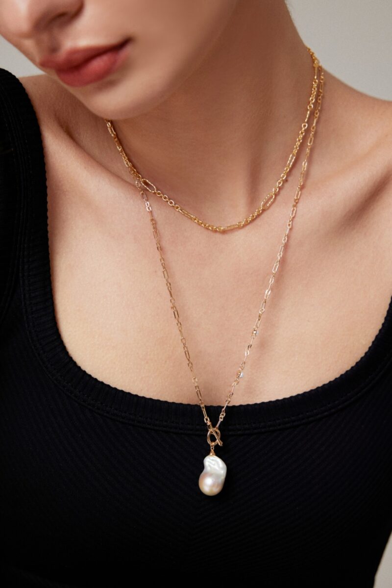 Close-up of a woman wearing a black top and two necklaces, one with a pendant featuring a large freshwater pearl, set against a neutral background. Elegant pearl jewelry