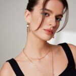 A portrait of a young woman with brown hair styled in a low bun, wearing a black sleeveless top, gold pearl jewelry earrings, and a layered necklace, gazing confidently at the camera.