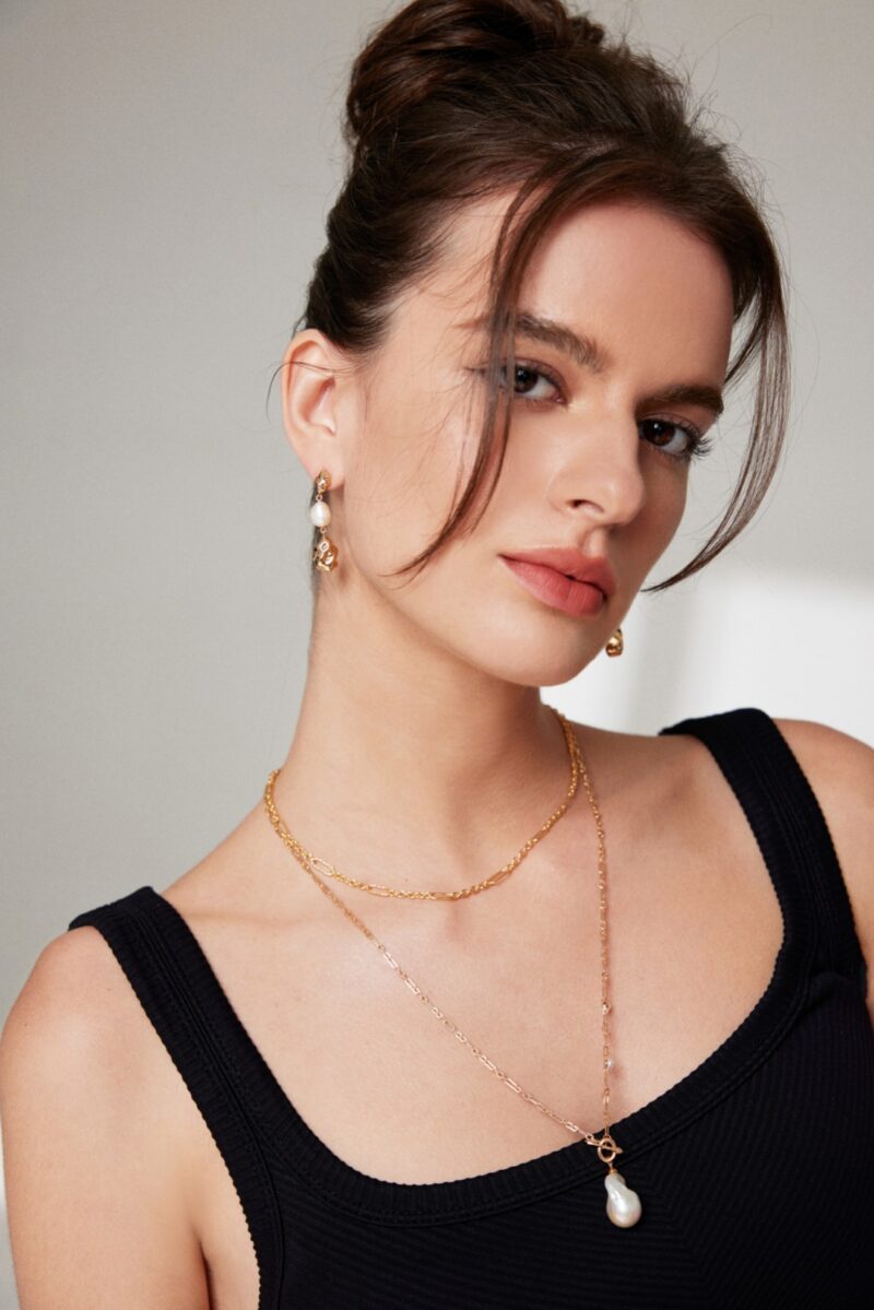 A portrait of a young woman with brown hair styled in a low bun, wearing a black sleeveless top, gold pearl jewelry earrings, and a layered necklace, gazing confidently at the camera.