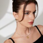 A woman with a delicate necklace and earrings looks thoughtfully at the camera, her hair styled in an elegant updo and her makeup featuring soft, natural tones. Elegant pearl jewelry