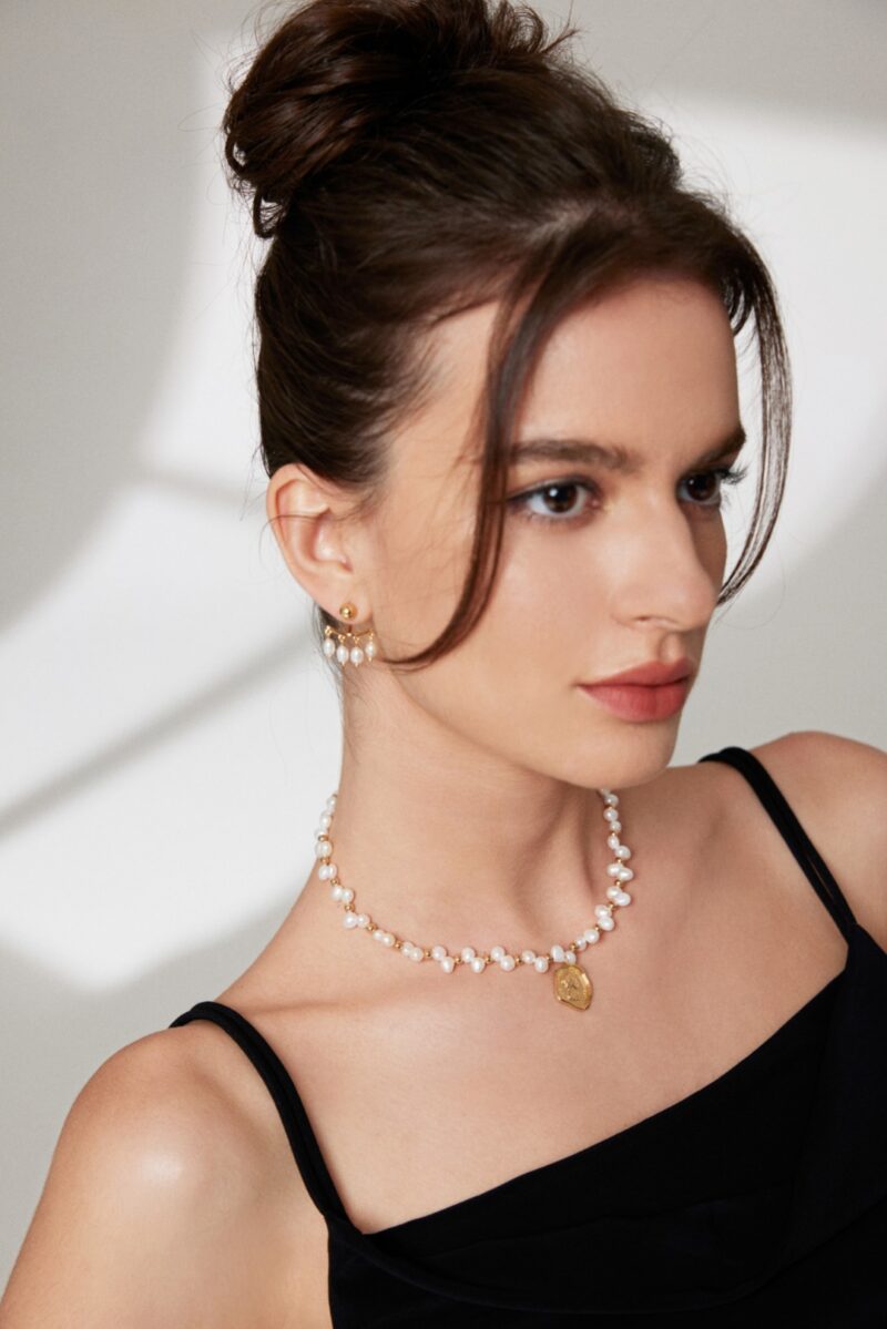 A woman with a delicate necklace and earrings looks thoughtfully at the camera, her hair styled in an elegant updo and her makeup featuring soft, natural tones. Elegant pearl jewelry