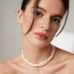 Close-up of a woman with an elegant hairstyle, wearing a necklace and looking at the camera. Her makeup features subtle eyeshadow and coral lipstick. Elegant pearl jewelry