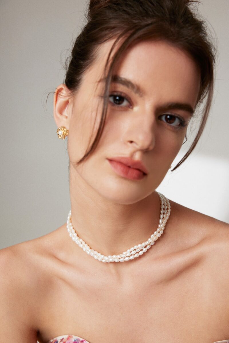 Close-up of a woman with an elegant hairstyle, wearing a necklace and looking at the camera. Her makeup features subtle eyeshadow and coral lipstick. Elegant pearl jewelry