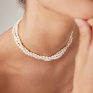 Close-up of a woman adorned with a necklace and a floral earring, highlighting elegant details against her skin. Her fingers touch her chin delicately. Elegant pearl jewelry