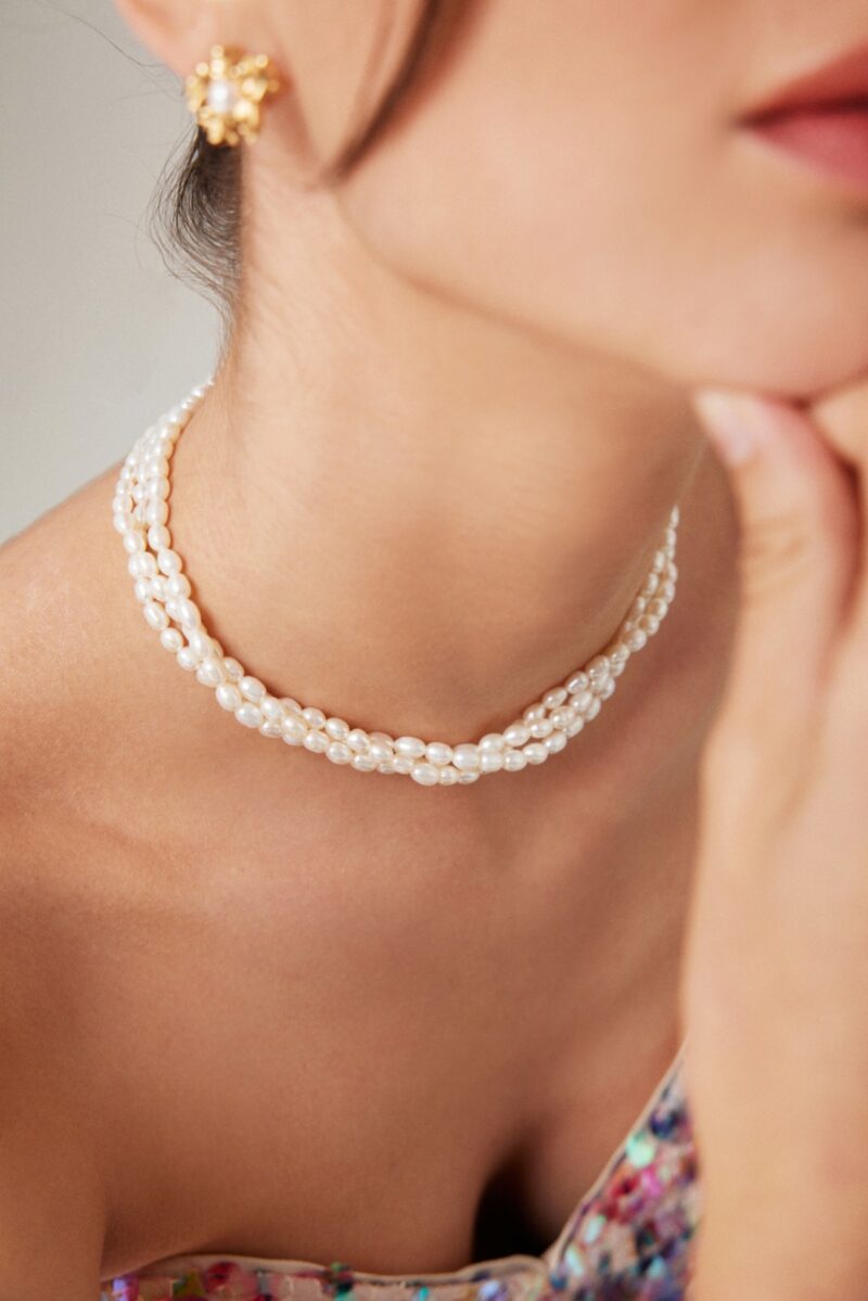 Close-up of a woman adorned with a necklace and a floral earring, highlighting elegant details against her skin. Her fingers touch her chin delicately. Elegant pearl jewelry