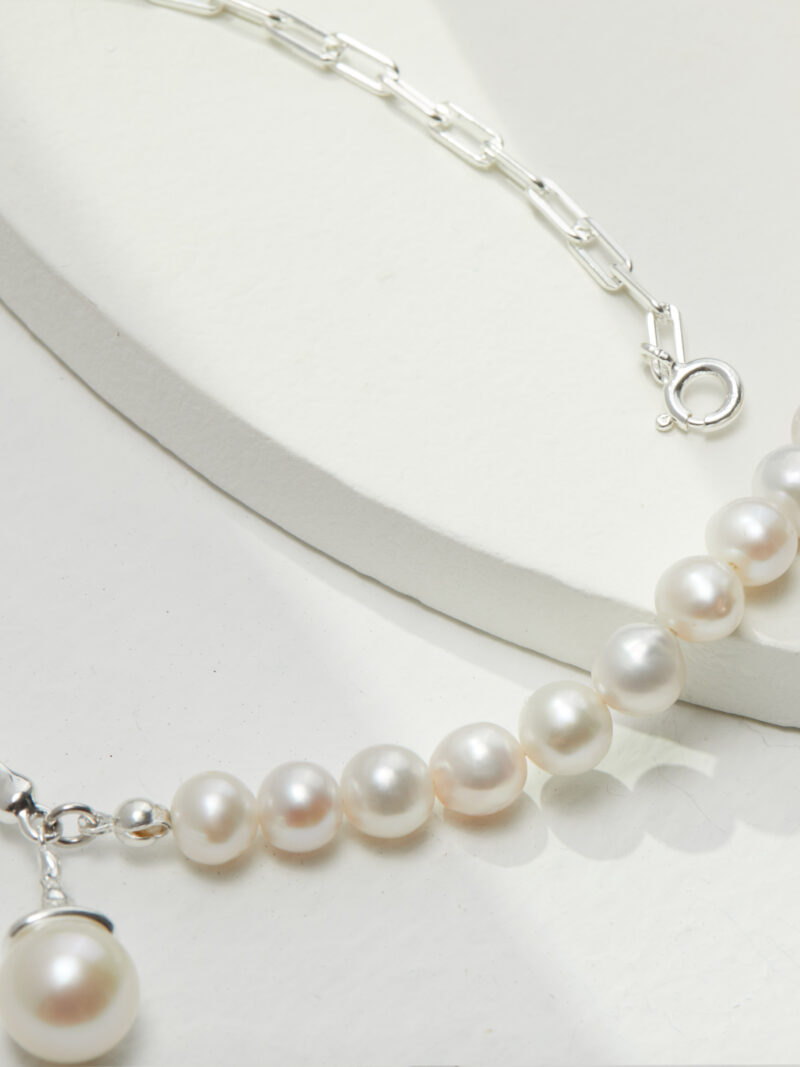 A close-up image of jewelry with a silver chain on a white display background, highlighting the lustrous pearls and the shiny metal clasp. Elegant pearl jewelry