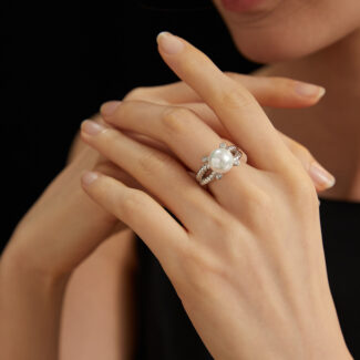 A close-up of a woman with elegantly manicured fingernails showcasing a large natural pearl ring, set against a black background. Only her crossed fingers and lips are visible.