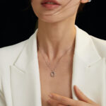 A close-up of a woman wearing a white blazer and a delicate necklace featuring the circular pendant, against a dark background. Her hair is windswept across her face. Elegant pearl jewelry