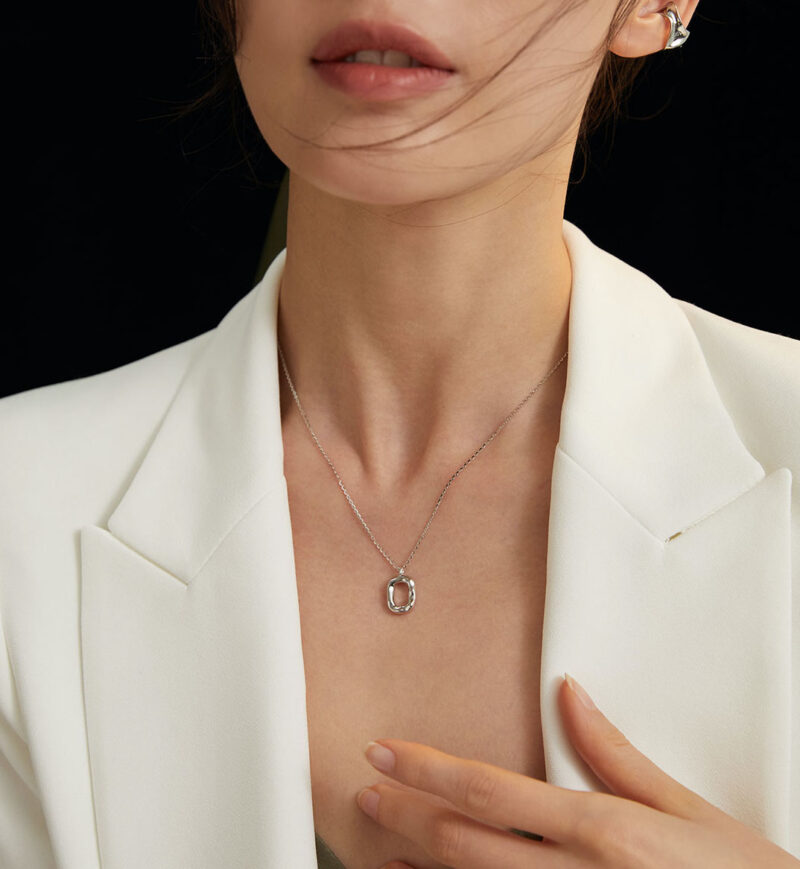 A close-up of a woman wearing a white blazer and a delicate necklace featuring the circular pendant, against a dark background. Her hair is windswept across her face. Elegant pearl jewelry