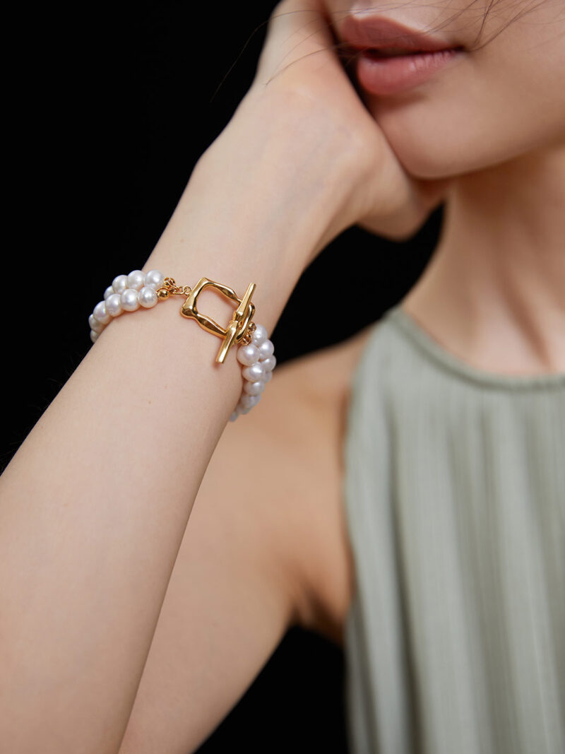 A woman wearing  jewelry on her wrist against a black background, her face partially visible and wearing a green top.