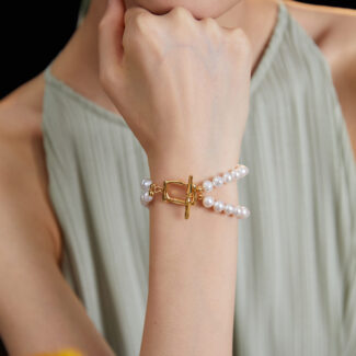 A close-up image of a woman's hand touching her chin, showcasing on her wrist. She wears a green pleated dress, and blurred orange flowers are visible in the foreground. Elegant pearl jewelry