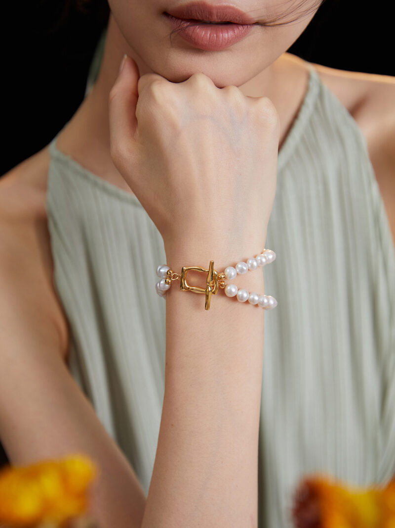 A close-up image of a woman's hand touching her chin, showcasing on her wrist. She wears a green pleated dress, and blurred orange flowers are visible in the foreground. Elegant pearl jewelry