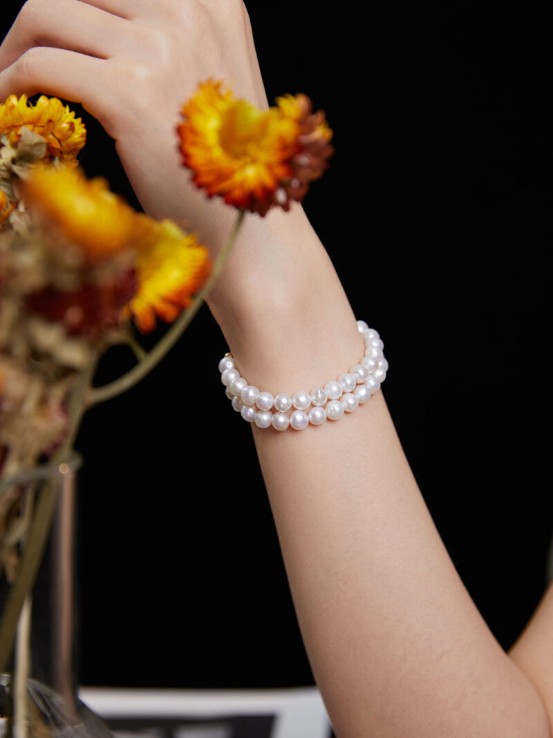 A woman's wrist adorned with a  bracelet, reaching towards orange and yellow flowers in a vase against a dark background.