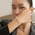 A close-up portrait of a woman showcasing a gold pearl bracelet and a necklace. She has sleek black hair pulled back, bold eyeliner, and is dressed in a black leather outfit, holding her chin