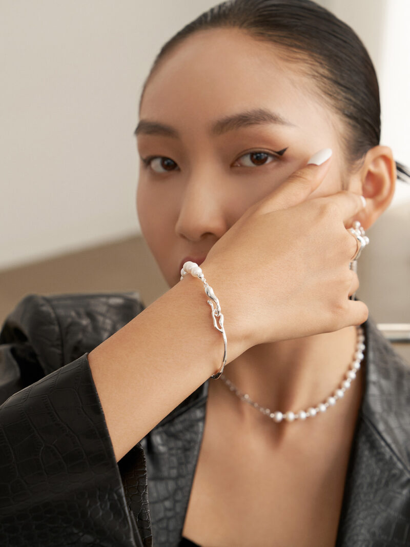 A close-up portrait of a woman showcasing a gold pearl bracelet and a necklace. She has sleek black hair pulled back, bold eyeliner, and is dressed in a black leather outfit, holding her chin