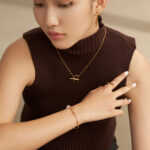 A woman in a brown sleeveless turtleneck and black pants, wearing product name jewelry, touches her chin thoughtfully, looking downwards. Elegant pearl jewelry