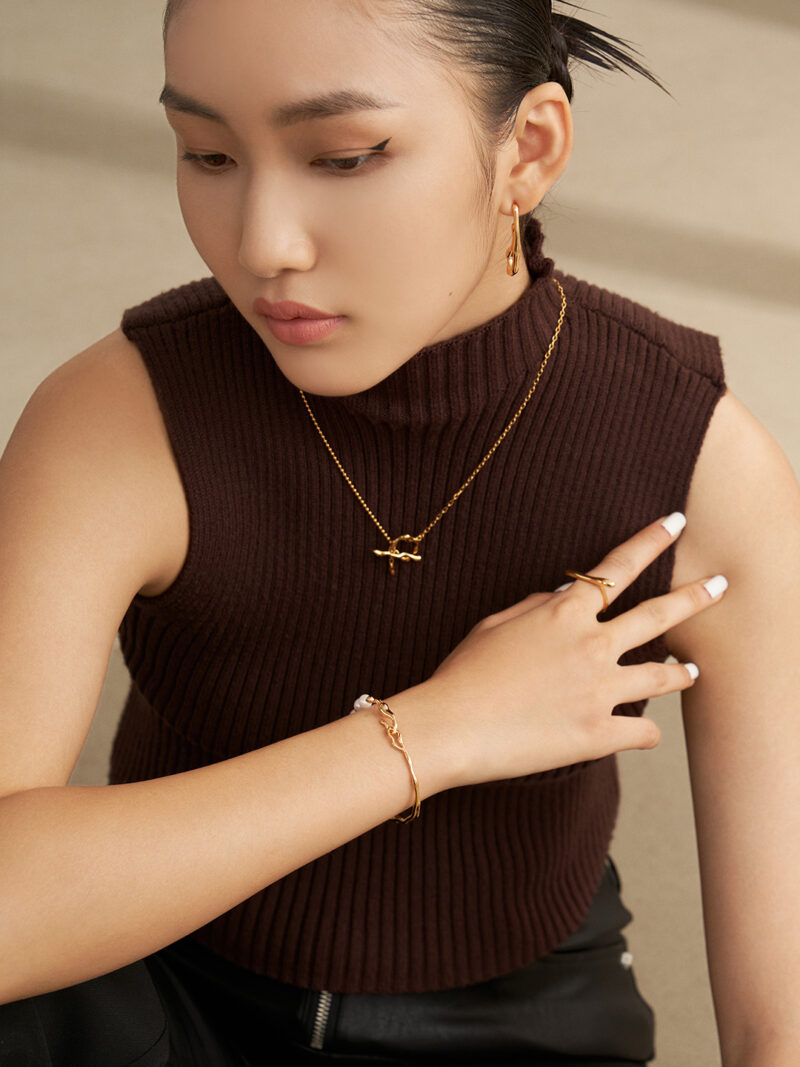 A woman in a brown sleeveless turtleneck and black pants, wearing product name jewelry, touches her chin thoughtfully, looking downwards. Elegant pearl jewelry