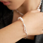 Close-up of a woman's wrist adorned with a bracelet, touching her shoulder, focusing on the elegant jewelry against a dark background. Elegant pearl jewelry