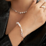 Close-up of a woman's neck and hand adorned with elegant jewelry, featuring a natural pearl necklace and a matching pearl bracelet with a sterling silver chain, against a textured black jacket.