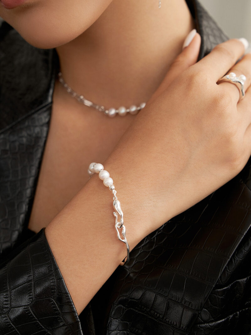 Close-up of a woman's neck and hand adorned with elegant jewelry, featuring a natural pearl necklace and a matching pearl bracelet with a sterling silver chain, against a textured black jacket.