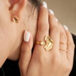 Close-up photo of a woman's hand with stylish white nail polish, showcasing unique gold rings with freshwater pearl and a small gold earring.