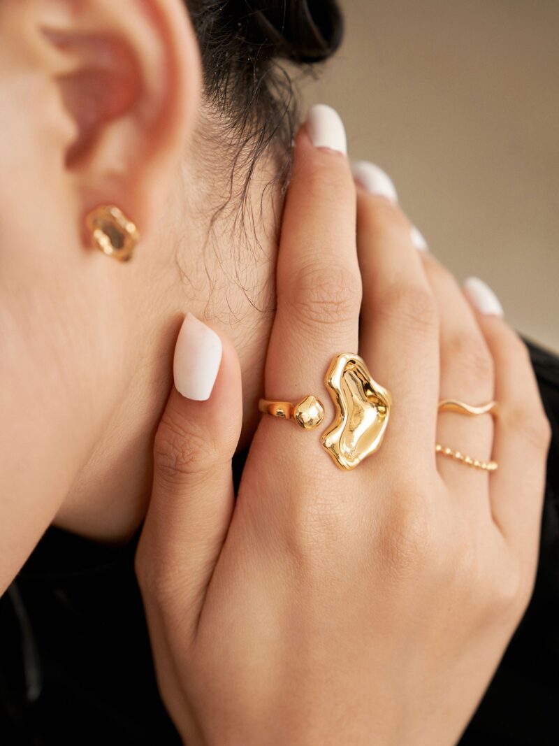 Close-up photo of a woman's hand with stylish white nail polish, showcasing unique gold rings with freshwater pearl and a small gold earring.