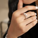 Close-up of a woman's hand with white manicured nails, touching her chin, wearing a unique silver ring shaped like a bird with pearl jewelry. Elegant pearl jewelry