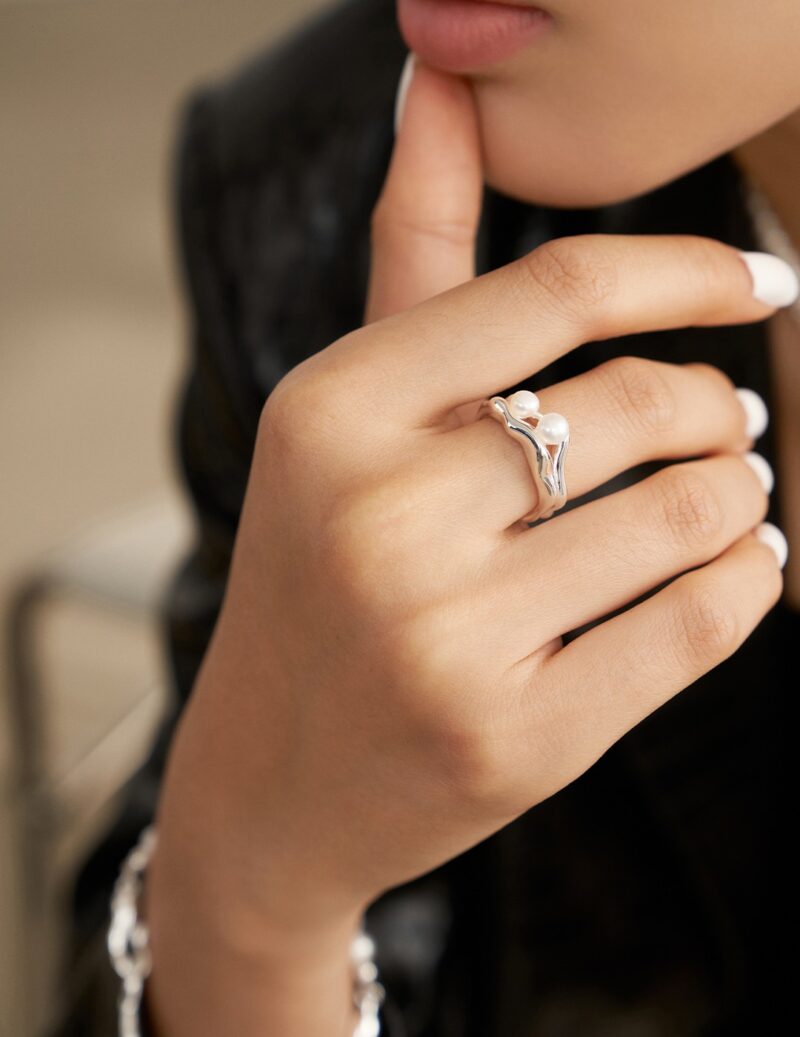 Close-up of a woman's hand with white manicured nails, touching her chin, wearing a unique silver ring shaped like a bird with pearl jewelry. Elegant pearl jewelry