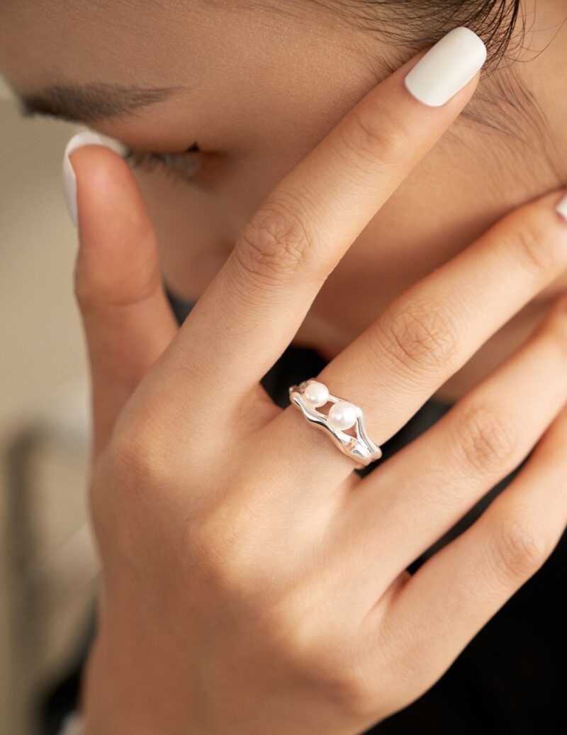 Close-up of a woman’s hand touching her face, showcasing a ring. She has manicured nails with white polish. Elegant pearl jewelry