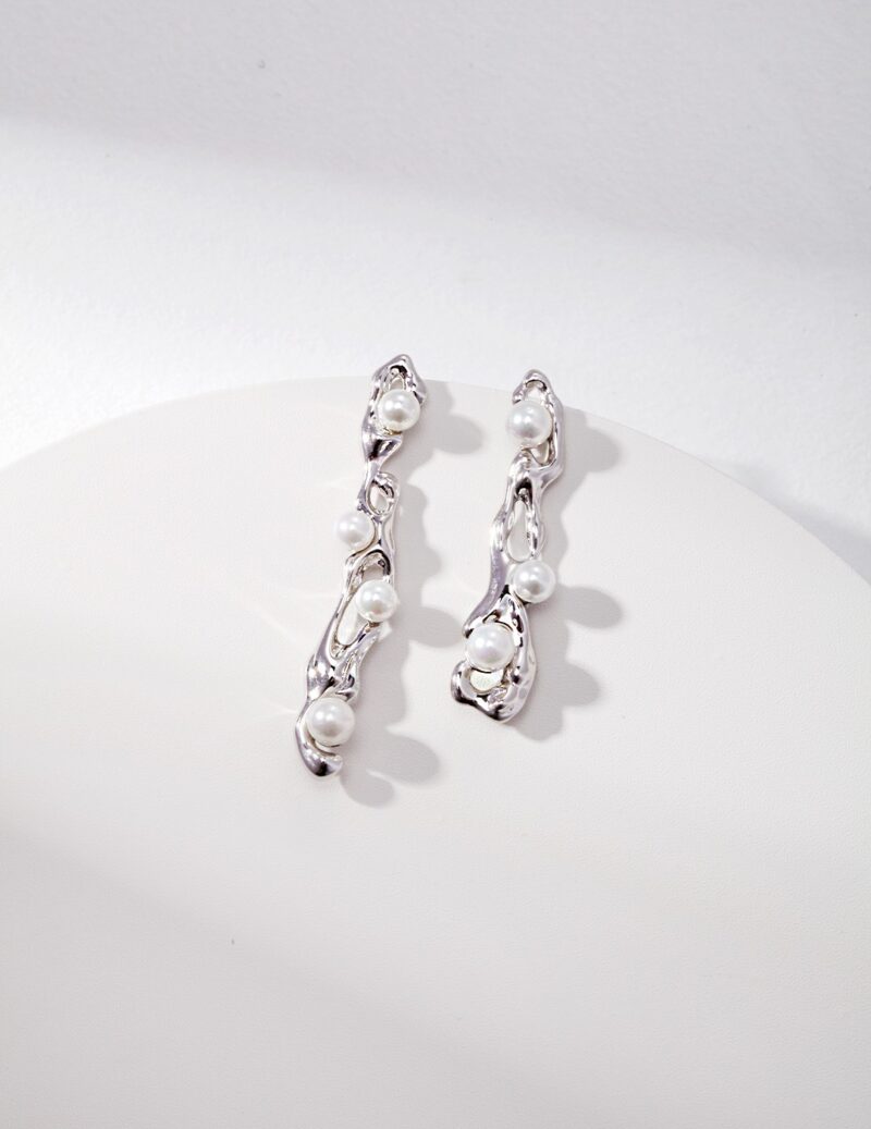 A pair of elegant earrings with a twisted design, featuring small natural pearls, displayed on a white circular surface against a white textured background. Elegant pearl jewelry