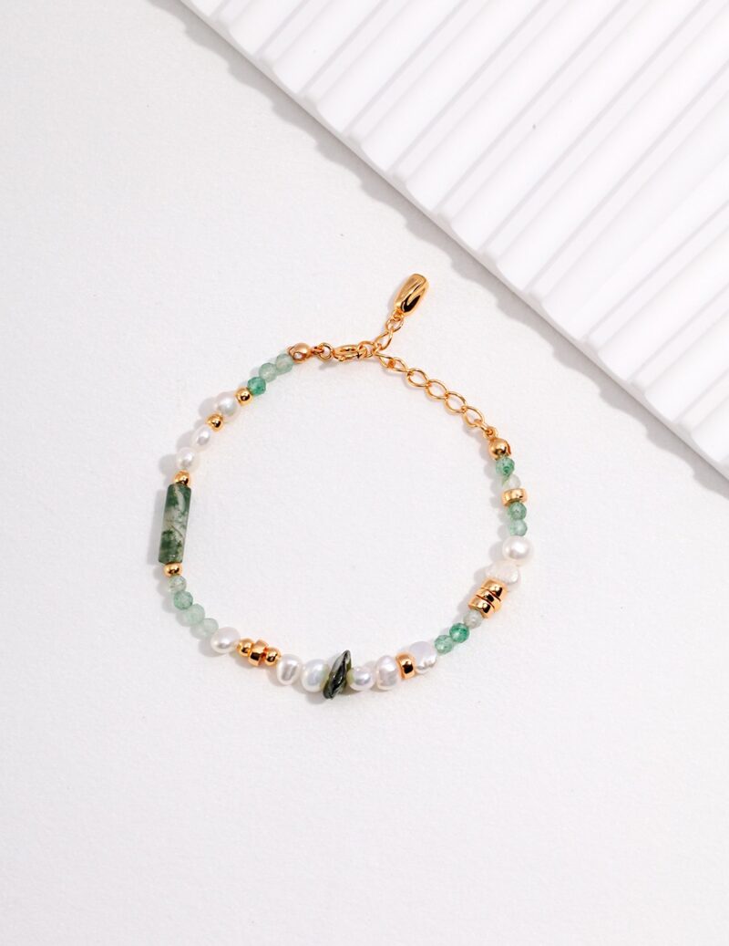 A delicate bracelet crafted with freshwater pearls, green beads, and golden clasps, displayed on a light gray surface beside a white ridged object. Elegant pearl jewelry