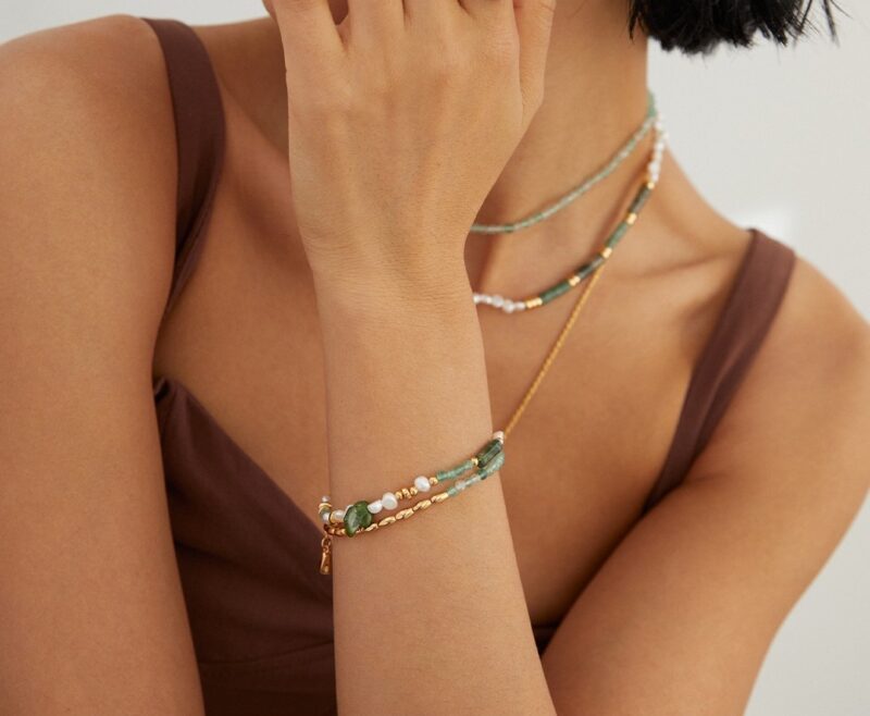 A woman in a brown top touches her neck, showing off a delicate as with green and white beads featuring freshwater pearl. Her face is partially visible, focusing on the jewelry. Elegant pearl jewelry