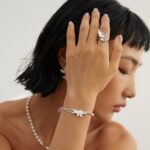 A woman showcases elegant jewelry, including rings and a bracelet with floral details, while gently touching her forehead with her hand adorned in the pieces. She has a short black hairstyle and wears pearl jewelry. Elegant pearl jewelry