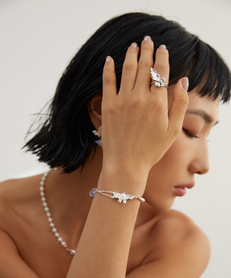 A woman showcases elegant jewelry, including rings and a bracelet with floral details, while gently touching her forehead with her hand adorned in the pieces. She has a short black hairstyle and wears pearl jewelry. Elegant pearl jewelry