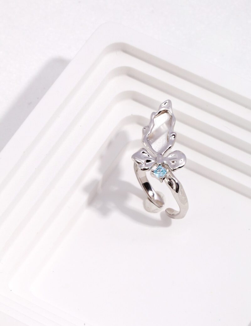 A butterfly-shaped  ring with a small natural pearl in the center, displayed on a white, grooved background. Elegant pearl jewelry