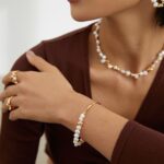 A close-up of a woman wearing elegant pearl jewelry including earrings, a necklace, and a bracelet. She is dressed in a brown top and touches her wrist gracefully. Elegant pearl jewelry