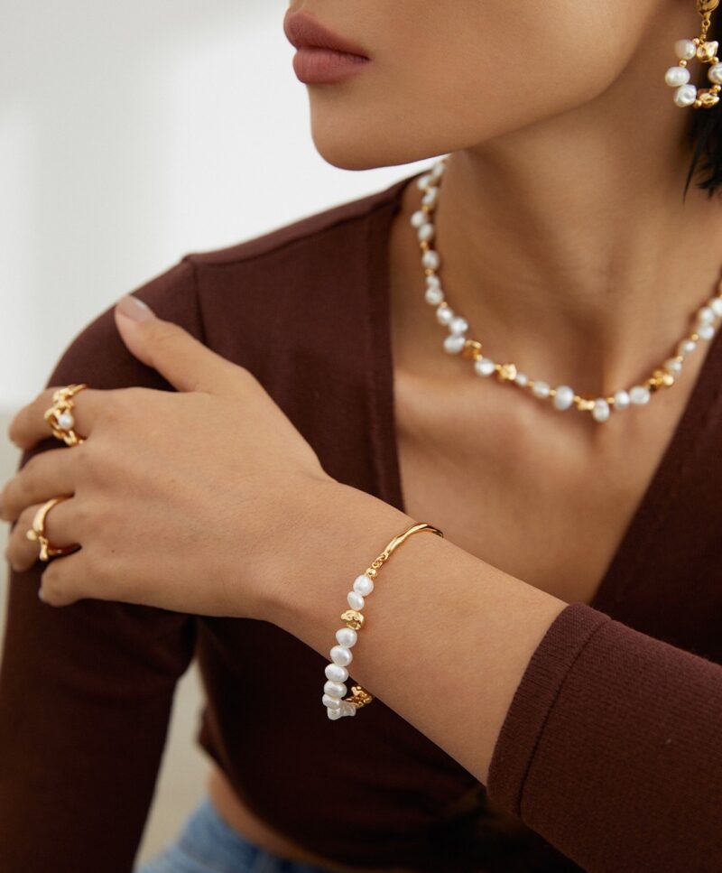 A close-up of a woman wearing elegant pearl jewelry including earrings, a necklace, and a bracelet. She is dressed in a brown top and touches her wrist gracefully. Elegant pearl jewelry